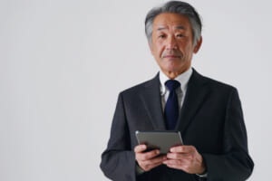 Content mature Japanese businessman using a digital tablet, isolated on a white background. A middle-aged CEO executive in a suit, an older male professional entrepreneur, looking at the camera.
